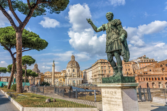 Statue of Julius Caesar at the Imperial Forums in Rome, Italy, with Trajan&rsquo;s Column, historic churches, and ancient ruins in the background under a bright blue sky.