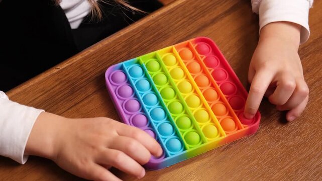 Bright rainbow push pop fidget lying on desk under small hands, trendy gadget for keeping children focused and calm, marketing for toy brands, e commerce listings and parenting blogs