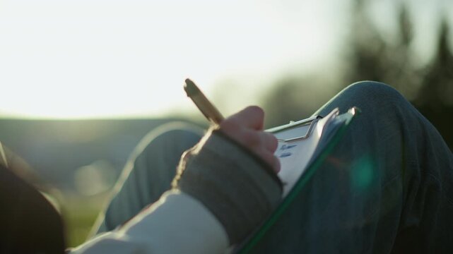 female artist creatively sketching outside, serene woman illustrating nature scenes outdoors during sunset, relaxed female illustrator capturing landscape on grassy hillside at dusk