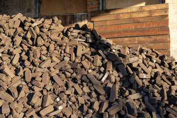 A large pile of dark bricks is placed next to a wooden wall, illuminated by sunlight. The scene captures a busy construction site environment