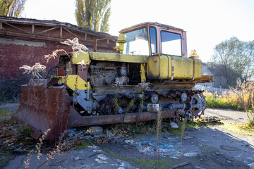 Fototapeta premium A yellow bulldozer sits rusting in a derelict industrial area, surrounded by overgrown weeds and remnants of an old building in the morning sun
