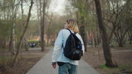 rear view white walker with backpack on paved park path, steady stride past trees and quiet benches, reflective commuter heading through woodland fringe toward city, muted spring tones