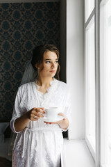 The bride in a white robe stands by the window holding a cup of coffee. She smiles softly, enjoying a calm moment before the wedding. A warm and peaceful morning atmosphere in a cozy room.