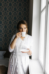 The bride in a white robe stands by the window holding a cup of coffee. She smiles softly, enjoying a calm moment before the wedding. A warm and peaceful morning atmosphere in a cozy room.