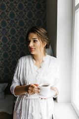 The bride in a white robe stands by the window holding a cup of coffee. She smiles softly, enjoying a calm moment before the wedding. A warm and peaceful morning atmosphere in a cozy room.