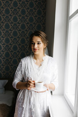 The bride in a white robe stands by the window holding a cup of coffee. She smiles softly, enjoying a calm moment before the wedding. A warm and peaceful morning atmosphere in a cozy room.