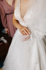The bride in a sparkling floral wedding dress holds her veil, showing her ring. A bridesmaid stands nearby. A delicate close-up capturing the beauty of wedding preparations and elegant bridal details