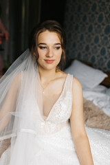 Portrait of a bride sitting indoors in her wedding dress. She looks at the camera with a calm, confident expression. Soft light highlights the lace details, veil, and floral bracelet, creating 