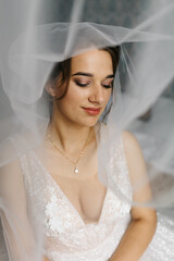 Bride smiling softly as her veil flows around her, creating a dreamy and romantic atmosphere. The close-up portrait highlights her makeup, lace wedding dress, and elegant jewelry in gentle natural 