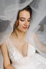 Bride smiling softly as her veil flows around her, creating a dreamy and romantic atmosphere. The close-up portrait highlights her makeup, lace wedding dress, and elegant jewelry in gentle natural 