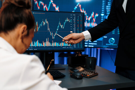 Smiling Asian business traders pointing to dynamic stock exchange data screen profit value online website program at night time at home office. Analyzing currency rate value on data report. Infobahn. - Powered by Adobe