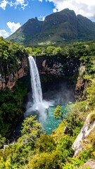 Towering waterfall cascades into a blue pool, surrounded by lush green trees and a majestic mountain backdrop