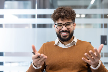 Happy male professional smiling at camera, sitting in a contemporary office setting with blurred glass partitions, gesturing while participating in an online meeting or interview