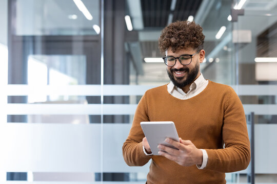 Young professional man in a contemporary office smiling as he uses a tablet to review business data and communicate online, reflecting tech-savvy, productive work life - Powered by Adobe