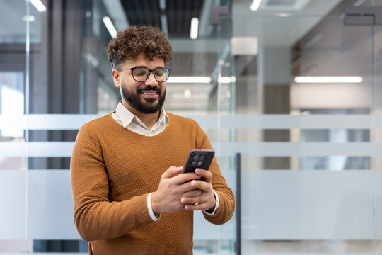 Young bearded man with curly hair smiling as he uses his smartphone in a modern office, conveying connectivity, mobile technology and professional, relaxed communication - Powered by Adobe