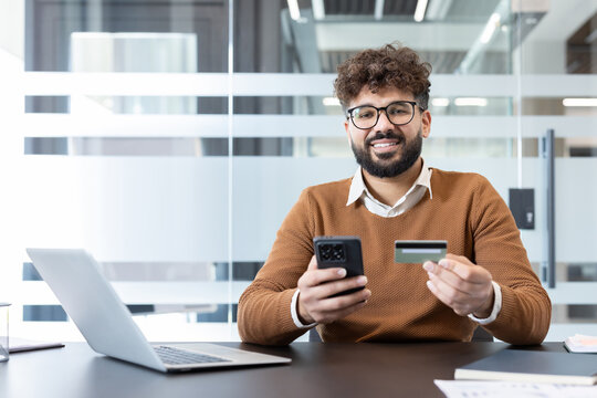 Young man smiling at camera holding a smartphone and a credit card, confidently making a secure online payment or transaction in a modern office environment
