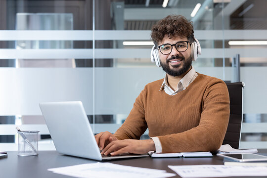 Young professional with curly hair, beard and glasses wearing white headphones at a desk, smiling at camera while working on a laptop in a bright modern office environment - Powered by Adobe