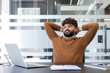 Young businessman leans back at his office desk, hands behind his head, smiling in a calm moment of satisfaction after finishing tasks, enjoying a peaceful work break