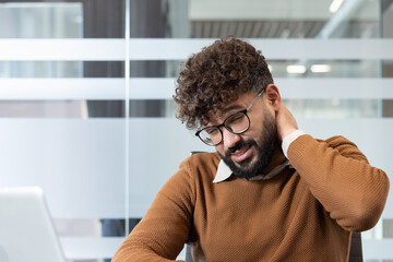 Young man experiencing neck pain and discomfort, touching his sore neck while working in a modern office, symbolizing stress, sedentary lifestyle, and health issues from long hours