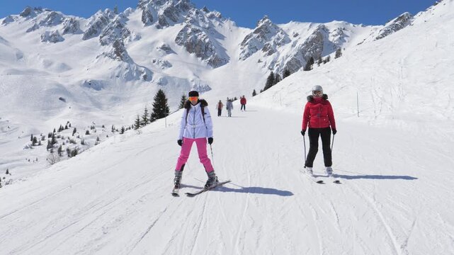 Women learning to ski together. Friends practicing parallel turns on snow. Bright blue sky over snow capped alpine slopes. Two women in colorful gear enjoy skiing among majestic mountain peaks