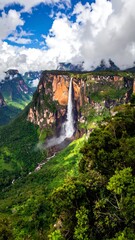 Fototapeta premium Tall waterfall cascading down a cliff face, surrounded by lush green foliage under a cloudy sky