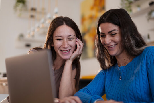 Two young women share a laugh while looking at a laptop screen together, enjoying a moment of connection and shared amusement.