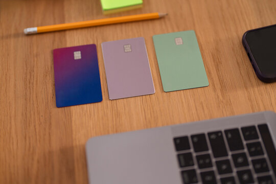 A person's hand reaches for a laptop, with credit cards, a phone, and a pencil on a wooden desk, suggesting financial planning or online transactions.