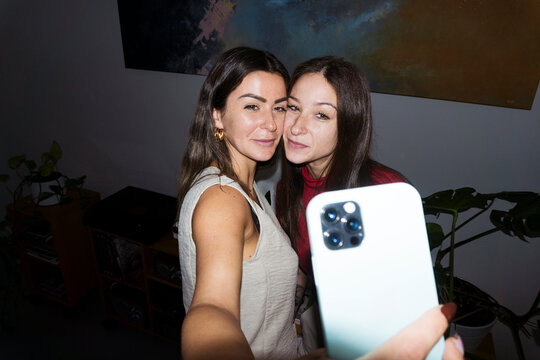 Two young women pose for a selfie with a smartphone in a dimly lit room, with plants and artwork in the background.