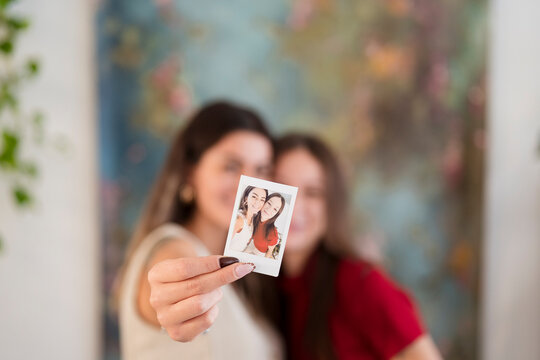 A woman holds up a small instant photograph of two smiling young women, with another woman blurred in the background.