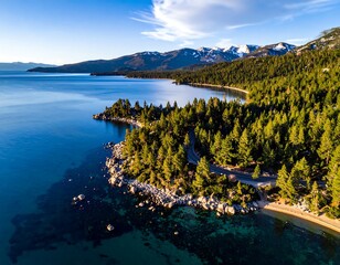 Lake's edge with rocky shoreline, dense forest, and snow-capped mountains in the background, under a clear blue sky