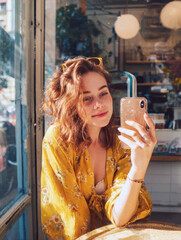 woman taking self-portrait on her smartphone inside cafe window, wearing yellow top and sunglasses, capturing casual, authentic moment reflecting trendy, contemporary, social media lifestyle