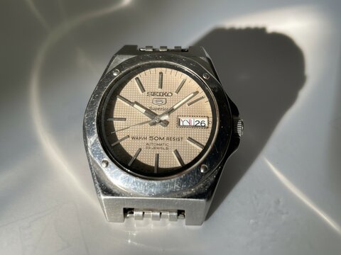 An old Seiko watch with a beige dial is displayed on a white background with strong shadows. This vintage timepiece features an automatic movement and a stainless steel case.