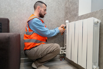 Worker specialist spends an hour repairing a radiator in a stylish living room. He wears a safety vest and focuses on fixing the heating system near the wall