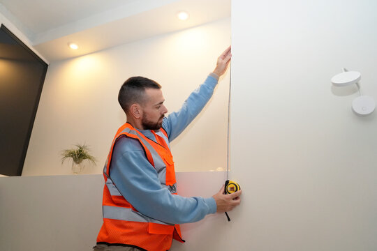 Worker specialist is measuring a wall with a tape measure to assist with home repairs in a well-lit living space. He is wearing an orange vest and focused on the task