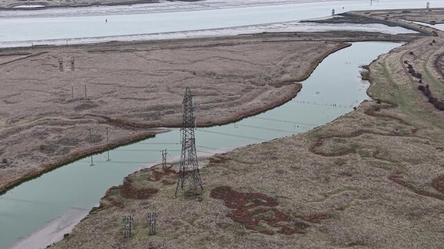 Electric power transmission tower next to the river. Aerial circular view of high voltage transmission cables running above the water. Marshes with bids on a grey cloudy day.