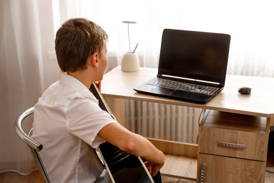 Rear view of a teenager or young man in a white shirt sitting at a desk with a laptop and an acoustic guitar. A scene of online learning or a hobby at home. - Powered by Adobe