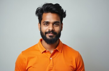 Fototapeta premium Young Indian man with beard and mustache in orange polo shirt. He looks serious and calm. Portrait of a man over grey background. Adult male with stylish hair and casual attire.