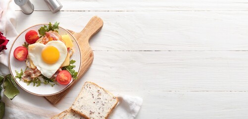 Plate with tasty heart-shaped fried egg, toasts, tomatoes, bacon and arugula on white wooden background with space for text