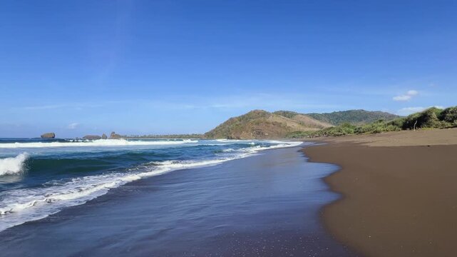 The sea in Jember, East Java. Indonesia. Watu Ulo beach with dark sand, mountains and coral rocks towering above the beach. The raging blue Indian ocean on a sunny day. 4К