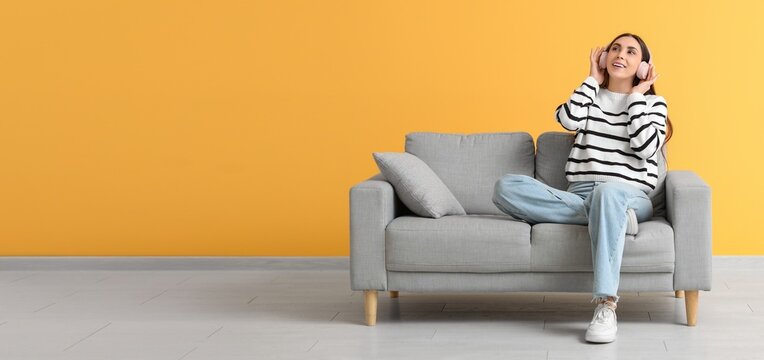 Happy woman in headphones sitting on comfortable sofa near yellow wall. Banner for design