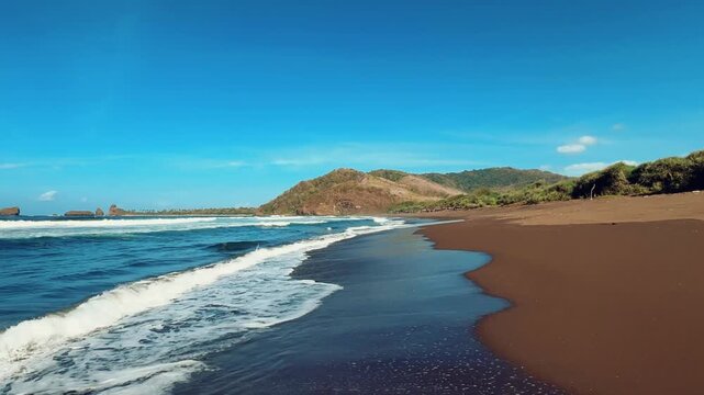 The sea in Jember, East Java. Indonesia. Watu Ulo beach with dark sand, mountains and coral rocks towering above the beach. The raging blue Indian ocean on a sunny day. 4К