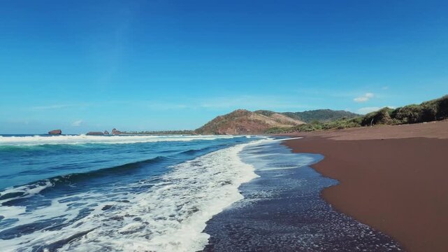 The sea in Jember, East Java. Indonesia. Watu Ulo beach with dark sand, mountains and coral rocks towering above the beach. The raging blue Indian ocean on a sunny day. 4К	