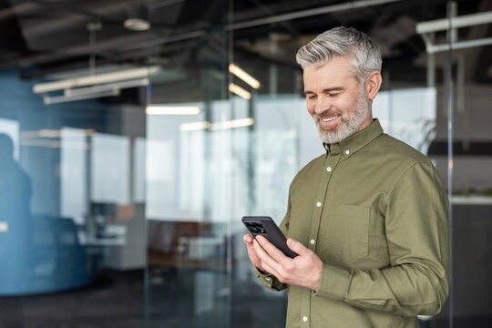 Mature businessman with gray beard smiling and using his smartphone while standing in a bright modern office, conveying connectivity, digital communication and professional confidence