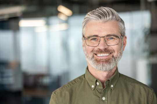 Happy professional mature man wearing eyeglasses and a casual shirt. Looking at camera while smiling widely in a modern office environment. Symbolizing success. Confidence. And achievement in business