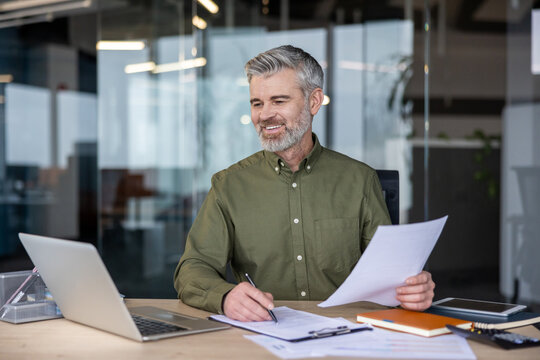 Happy mature businessman sitting at an office desk, smiling while reviewing financial paperwork and using a laptop, representing success and diligent work - Powered by Adobe