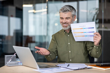 Mature businessman sitting at desk in modern office showing financial metrics report to laptop camera during online video call, discussing business strategy and results