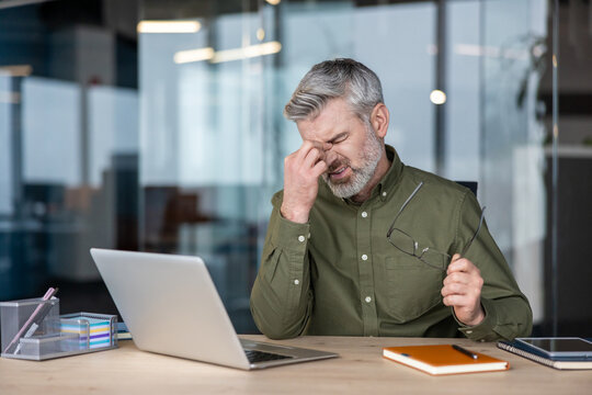 Mature man experiencing digital eye strain and a painful headache, rubbing his eyes while working on a laptop at his office desk, symbolizing workplace fatigue and burnout - Powered by Adobe