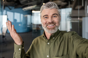Smiling mature businessman having a video conference call or recording a video, waving with a friendly gesture and looking directly at the camera in a modern office environment