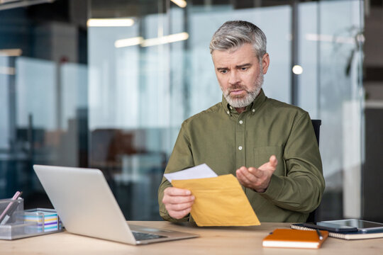 Mature businessman sitting at his desk, opening a document from an envelope and reacting with shock and confusion to unexpected bad news or financial problems