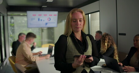 Smiling businesswoman extends horizontal smartphone with green screen toward camera after liking what she sees, diverse team working in office meeting background